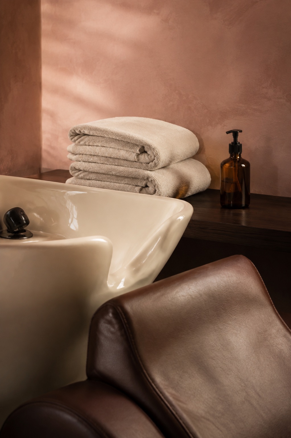 An ivory wash basin, folded cotton towels, and an amber pump bottle in the warm salon light