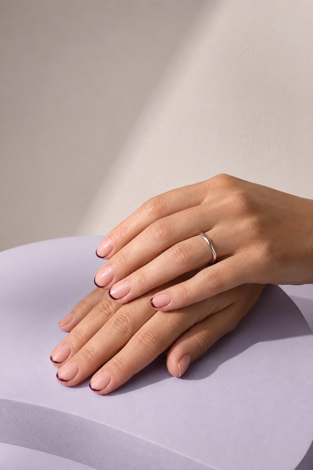 Hands with a sheer pink and deep plum French manicure resting on a lilac display plinth